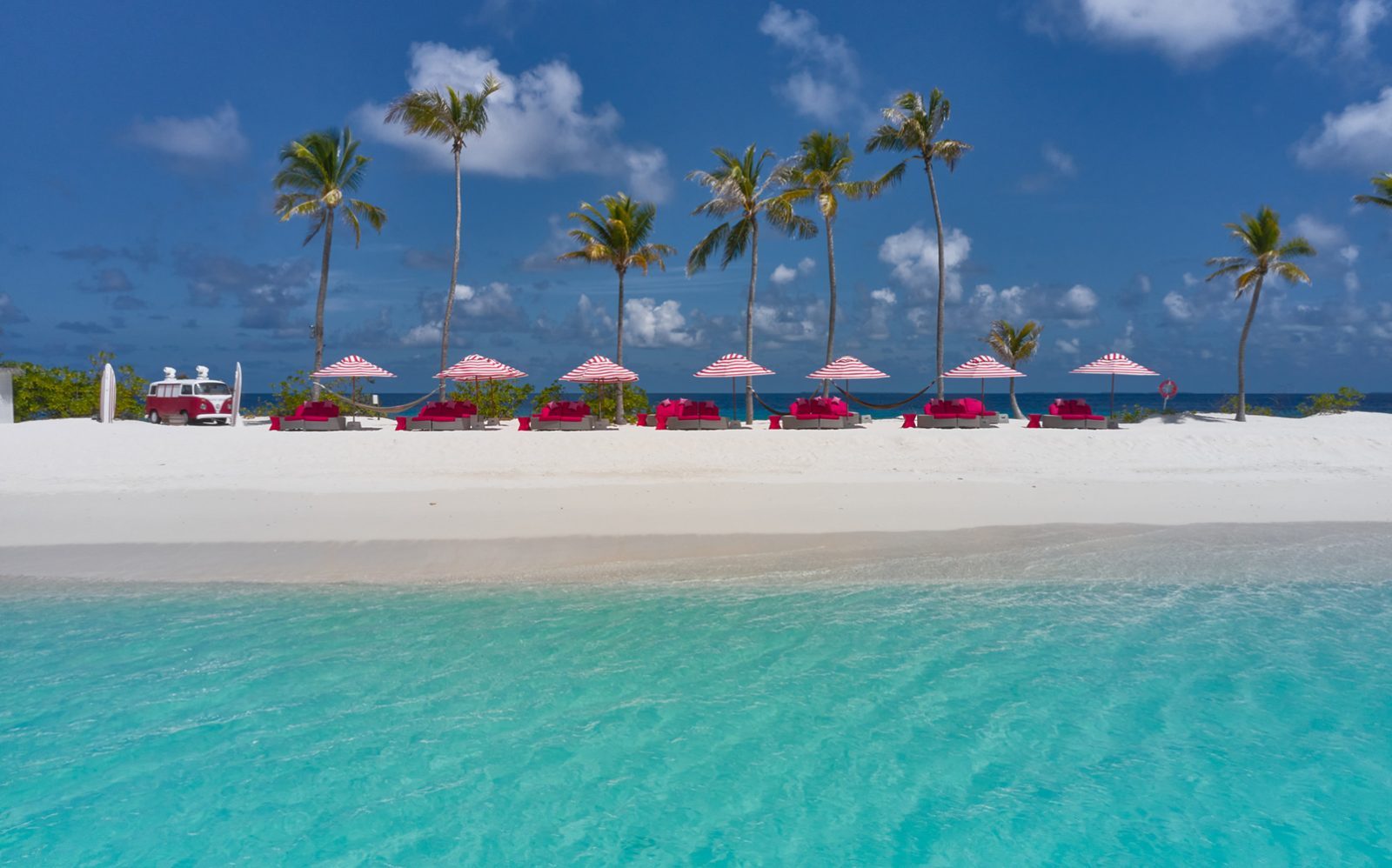 Weißer Sandstrand mit türkisblauem Wasser, Palmen, rote gestreifte Sonnenschirme und rote Lounge-Möbel; links ein VW-Transporter.