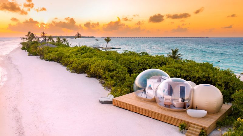 download (15)_quer Sonnenuntergang am Strand; drei glaskugelförmige Bubble-Pods auf Holzdeck am Meer, Palmen im Hintergrund.