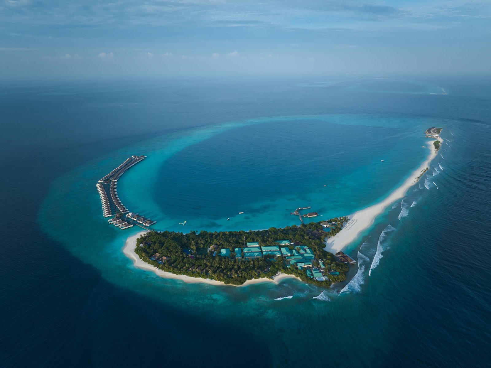 Luftaufnahme einer Tropeninsel mit Bungalows über dem Wasser, heller Sandstrand und türkisblauem Lagunenwasser.