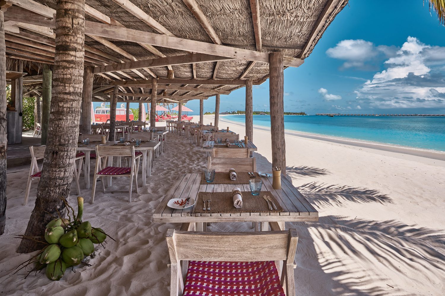 Open-Air Strandrestaurant aus Holz unter Strohdach, Tische am Sand, Blick aufs türkisblaues Meer