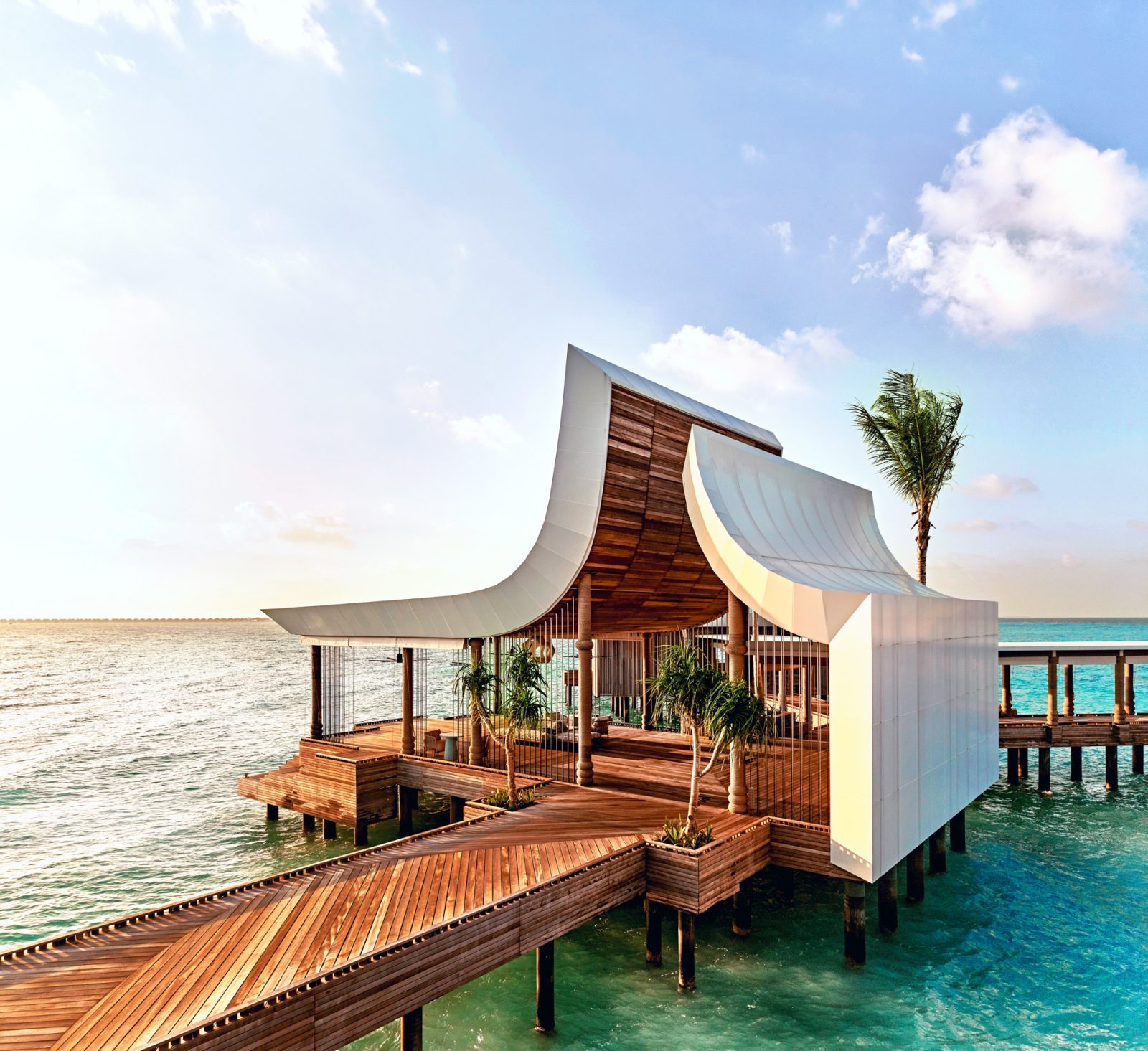 Architektonische Holzdecks am Meer mit geschwungenen weißen Dächern, Palmen und Wasser im Hintergrund