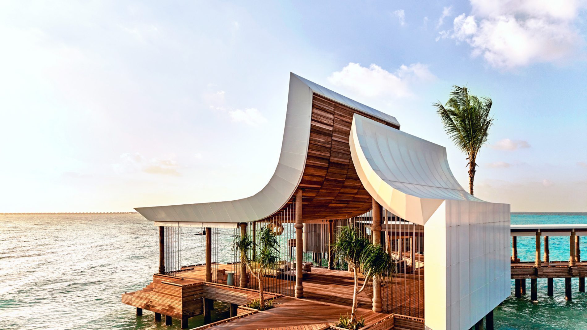 Architektonische Holzdecks am Meer mit geschwungenen weißen Dächern, Palmen und Wasser im Hintergrund