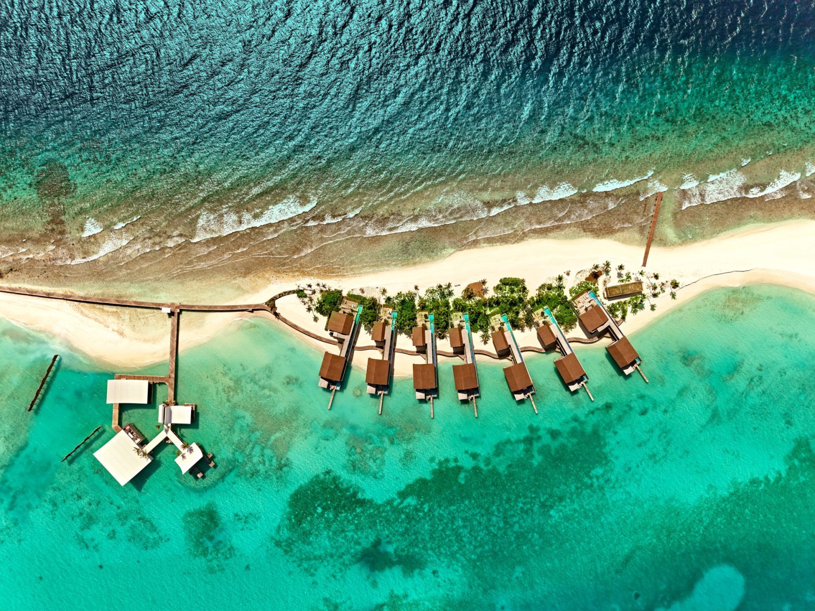 Luftaufnahme: türkisfarbenes Meer, Sandstrand, Bungalows auf Pfählen über dem Wasser, Stege, Palmen am Ufer.