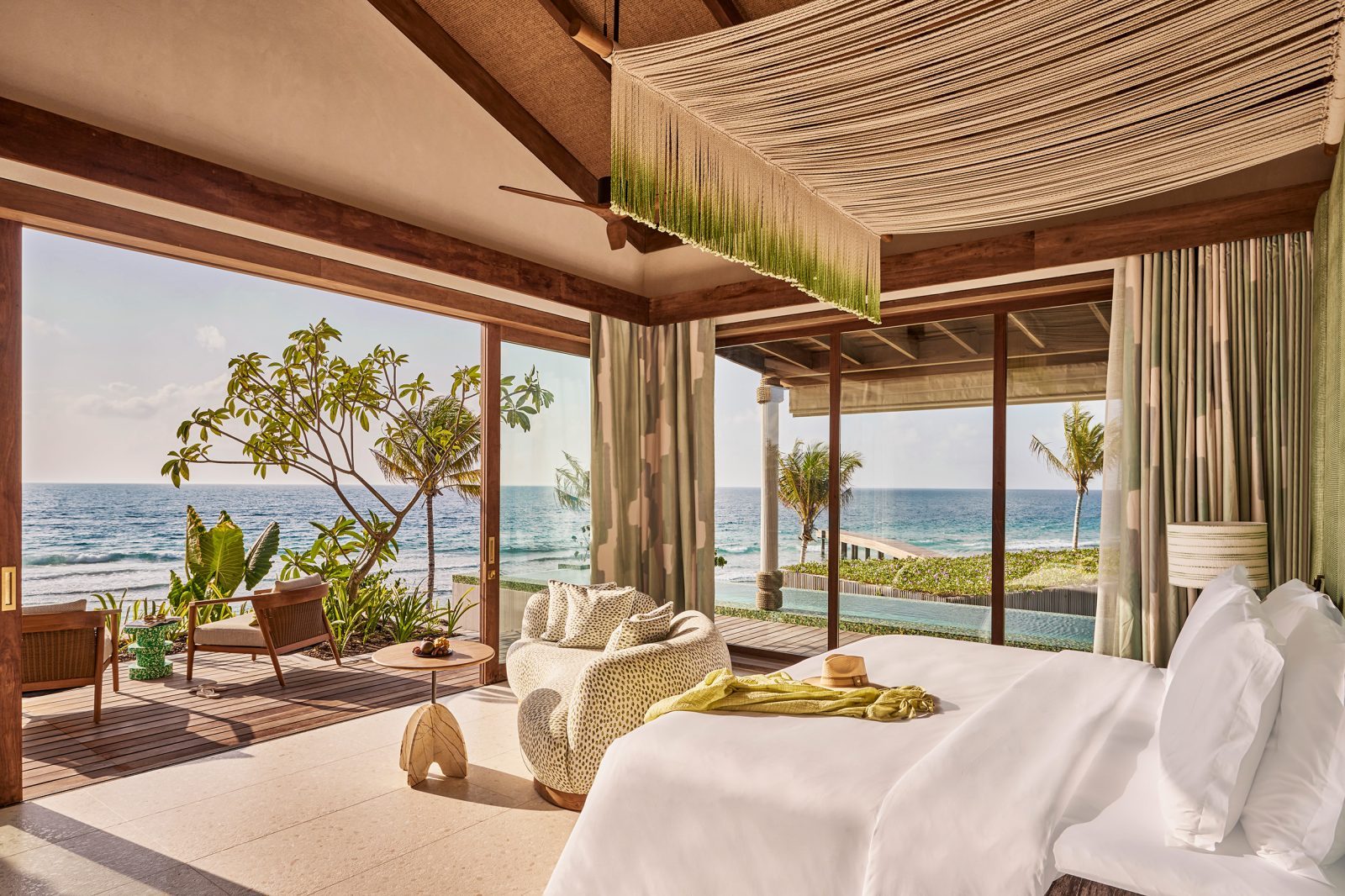 Schlafzimmer mit bodentiefen Glasfronten zum Strand, Meerblick, Lounge-Sessel, Pflanzen und Vorhänge.