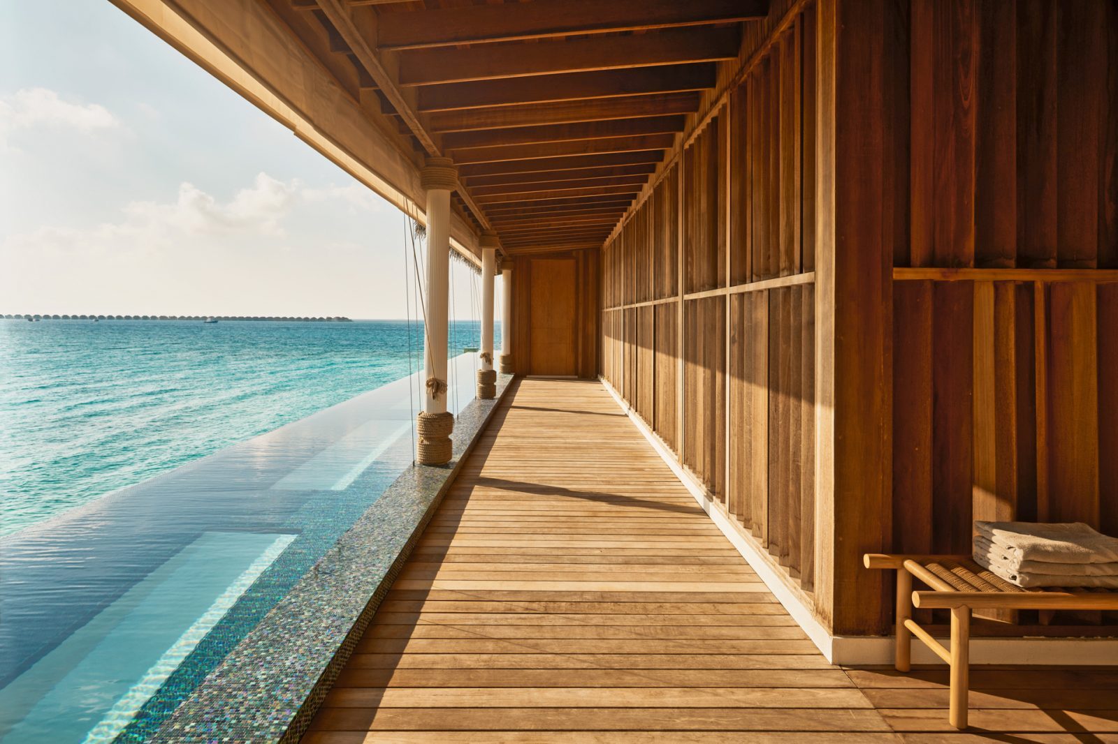 Villa mit Holzveranda am Meer: Blick auf Infinity-Pool links, ruhige Holzwand rechts, sonniges Ambiente.