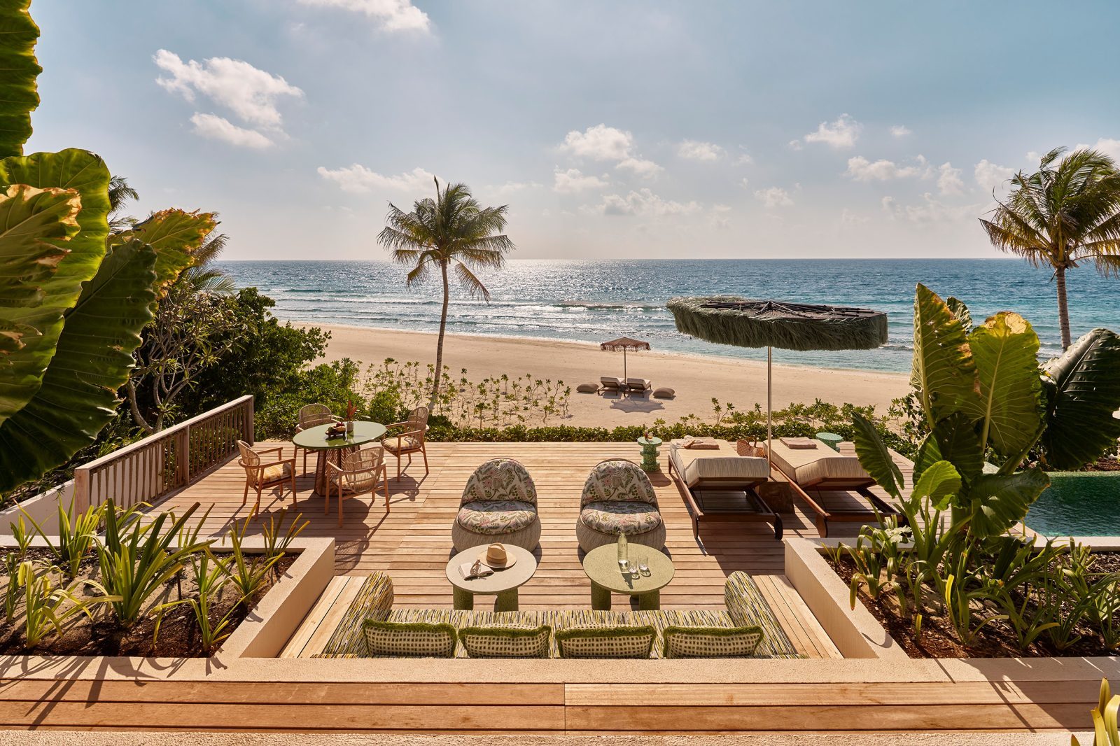 Terrasse am Strand mit Esstisch und Lounge-Sesseln, Palmenrahmen, Blick aufs Meer und Sonnenreflexe.
