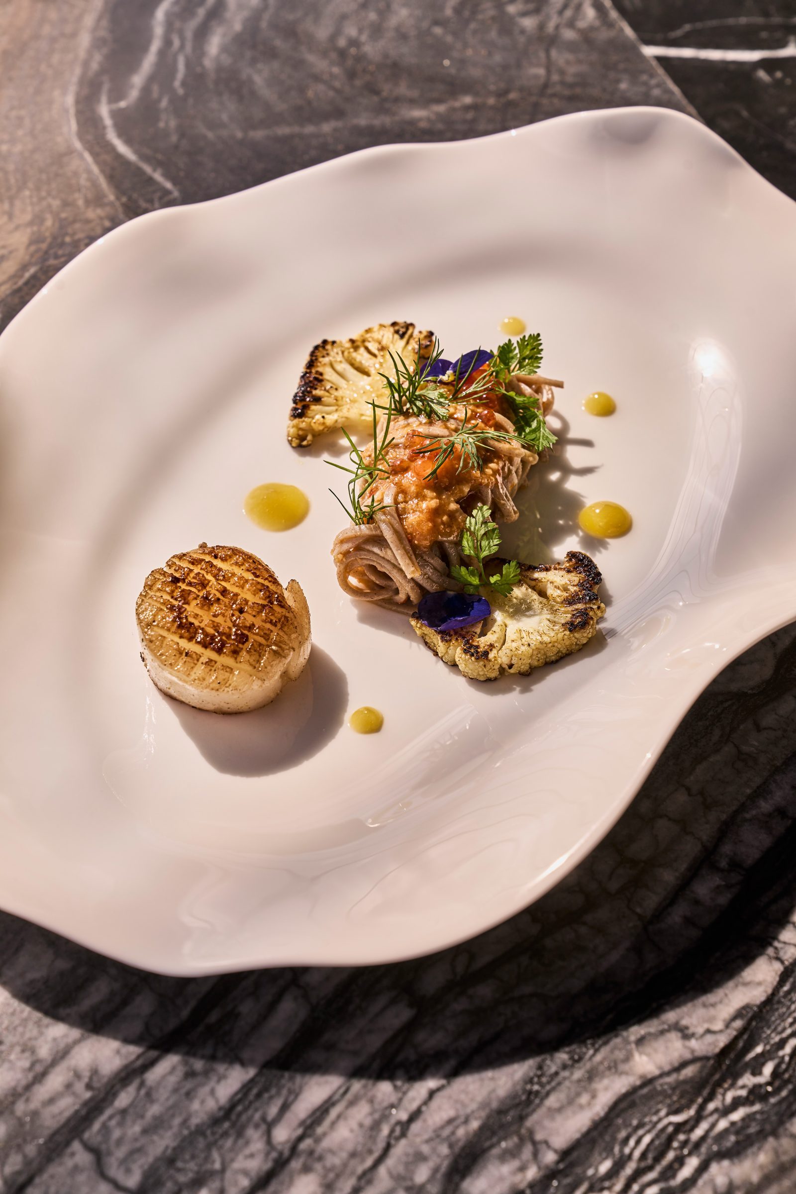 Gegrillte Blumenkohlröschen, Nudelnest, Kräuter, gelbe Tropfen-Sauce auf weißem Teller vor dunklem Stein.