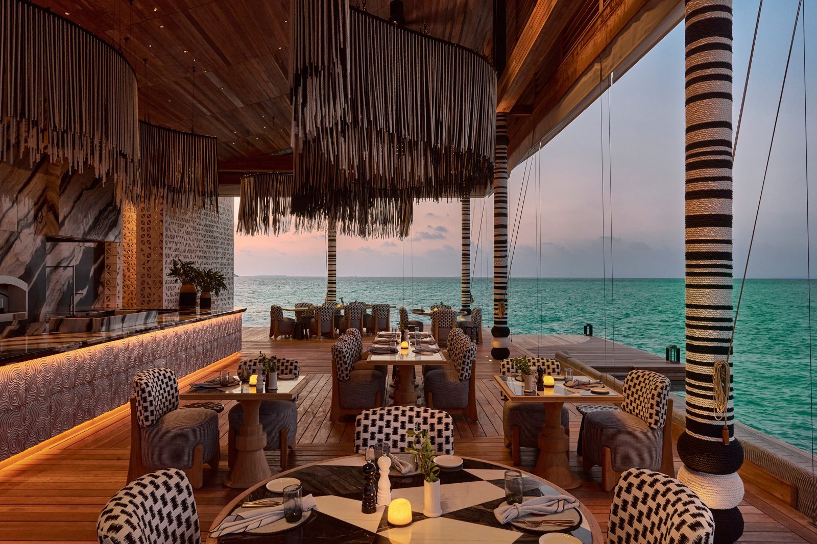 Offenes Strandrestaurant am Meer mit Holzboden, runden Tischen, schwarz-weiß gemusterten Stühlen und hängenden Dekorstreifen.