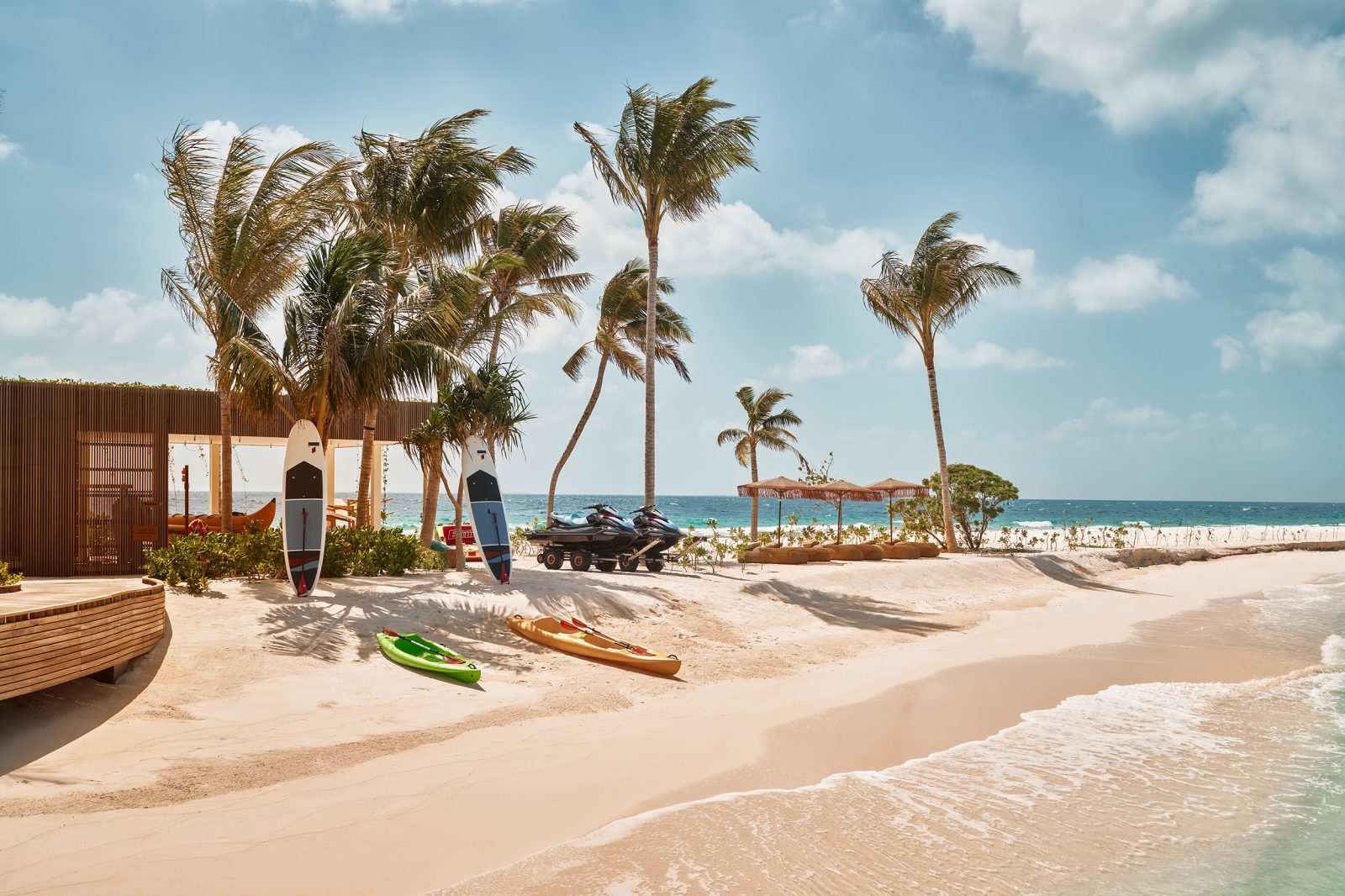 Strand mit Palmen, Surfbrett- und Jetski-Ausstattung, heller Sand, Meer und blauer Himmel.